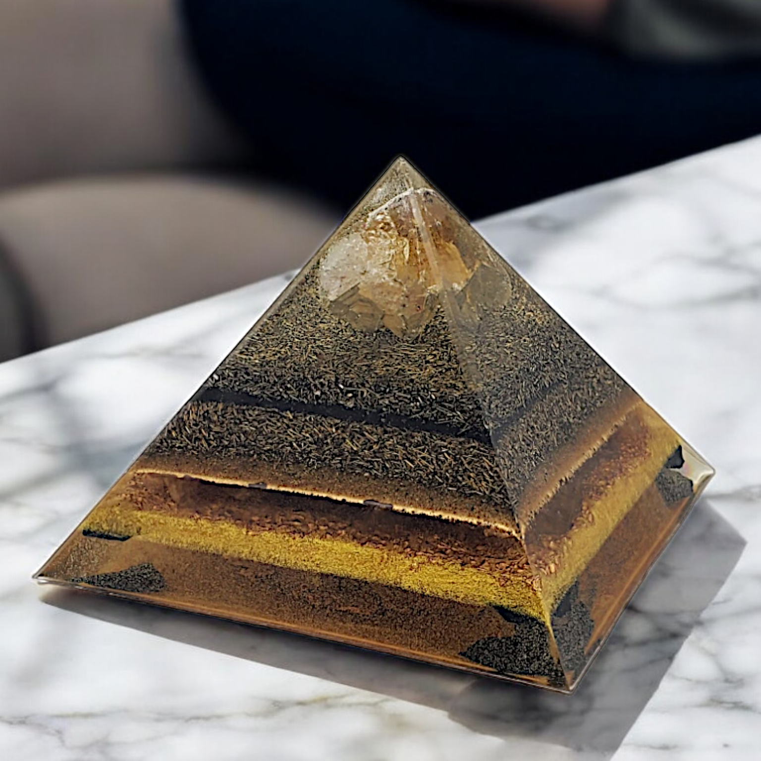 Pyramid-shaped orgonite with layered structure on a marble surface, blurred person in the background